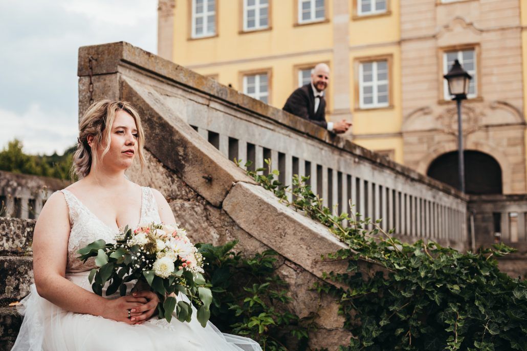 Braut mit Brautstrauß sitzt auf einer Treppe am Schloss. Bräutigam schaut über die Brüstung der Brücke